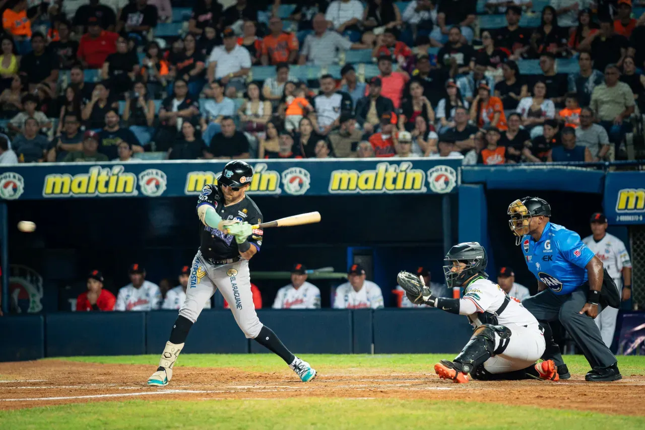 Bateador de Bravos de Margarita conecta la pelota frente al receptor de Águilas y el árbitro principal en Maracaibo.