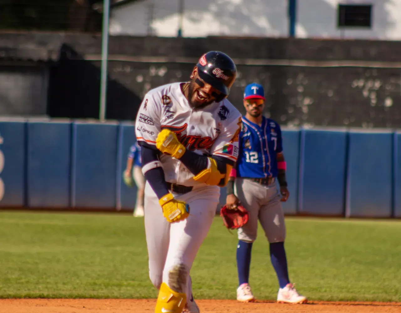 Pelotero de Tigres de Aragua, con uniforme blanco, casco y guantes amarillos, celebra sobre el terreno frente a un defensor de Tiburones.