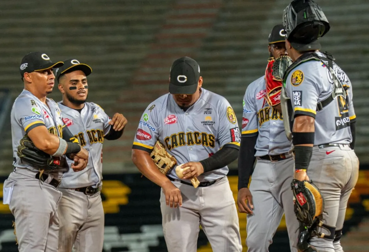 Peloteros de Leones del Caracas, con uniformes grises, se reúnen junto al montículo durante un encuentro.